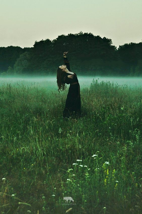 graceful women in dark dress in the middle of green field, that is coved in fog.